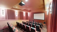 This purpose-built private screening room delivers a true cinematic experience in an intimate, well-appointed setting. The room is designed with dedicated theater-style seating arranged in tiered rows, ensuring clear sightlines to the large projection screen mounted at the front of the space. Each seat features a dark upholstered frame with pink cushioned armrests and folding sides, combining comfort with classic movie-house aesthetics.

A ceiling-mounted projector anchors the technical setup, providing high-quality image projection onto the wide screen. The walls are dressed in warm rose-toned drapery panels, contributing to both the acoustic performance and the rich, atmospheric ambiance of the room. Decorative wall sconces are evenly spaced along the side walls, offering subtle ambient lighting that enhances the theatrical feel without detracting from the viewing experience.

Wood paneling along the lower portion of the walls adds a layer of warmth and texture, while the ceiling features a coffered design with recessed lighting accents. The carpeted flooring further aids in sound absorption, ensuring an acoustically refined environment. Overall, this screening room is a thoughtfully designed space that balances functionality with comfort, making it ideal for private film screenings, presentations, or entertainment events.