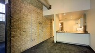 This single-floor space showcases a compelling blend of industrial and contemporary design. A dominant feature is the expansive exposed brick wall, which adds raw texture and warmth to the interior. Visible ductwork runs along the upper portion of the brick wall, reinforcing the industrial aesthetic. The flooring consists of polished concrete, providing a sleek, durable surface that complements the rougher textures of the brick. Recessed lighting is embedded in the smooth white ceiling, offering a clean contrast to the rugged wall surfaces. A kitchen or wet bar area is positioned to one side, featuring a dark stone countertop, a sink, and cabinetry. This area is finished with crisp white walls, creating a distinct visual separation from the brick-dominated main space. A passageway visible in the background, accented by pendant globe lighting, hints at additional areas beyond. The interplay between the weathered brick, polished concrete, exposed mechanical elements, and refined finishes gives the space a loft-like character that balances utilitarian origins with modern sensibility, making it well-suited for a variety of uses.