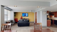 This single-floor space is thoughtfully designed to accommodate communal living and gathering. The open-plan layout seamlessly connects the living room, dining room, and kitchen, promoting a sense of openness and ease of movement throughout.

The living area is a welcoming focal point, featuring carpet tile flooring, large windows with horizontal blinds that allow ample natural light, and a bold yellow accent wall that anchors the entertainment zone. A wall-mounted flat-screen TV, a leather sofa, and eclectic seating — including a red tufted bench and a modern shell chair — create a relaxed, social atmosphere. Decorative green glass wall art adds a distinctive artistic touch.

The dining area accommodates multiple guests with two wooden tables and matching slatback chairs, complemented by a framed nature-themed artwork and a wall-mounted light fixture. Windows along the dining wall continue the theme of natural light throughout the space.

The kitchen is functional and well-appointed, featuring rich cherry wood cabinetry, light stone-finish countertops, and black appliances including a refrigerator, range, and dishwasher. A textured stone-look floor grounds the galley-style layout, which offers generous storage and counter space.

A hallway connects the main areas, completing this efficiently organized and inviting communal space.