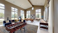 This generously proportioned game room is designed for social entertainment, distinguished by its dramatic vaulted ceilings with exposed beam accents and an abundance of natural light streaming through floor-to-ceiling windows. The large-pane windows line multiple walls, offering sweeping views of the surrounding landscaped grounds and water features, creating a bright and welcoming atmosphere throughout.

The space is well-equipped with a wide array of gaming options. A central billiards table serves as a focal point, complemented by a foosball table, a full-length shuffleboard table, and classic arcade machines. A large tic-tac-toe wall feature adds a playful decorative element. Cue stick wall racks are conveniently mounted near the pool table area.

A comfortable lounge seating area with colorful upholstered sofas and accent chairs provides a relaxed gathering spot, anchored by a patterned area rug over light hardwood-style flooring. A separate smaller seating nook near the windows offers a quieter retreat. The walls feature bold geometric color-block panels in teal, gold, and neutral tones, adding a vibrant, modern character to the overall design.
