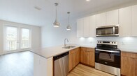 This single-floor unit presents a clean, contemporary design highlighted by an open-concept layout and consistent light hardwood flooring throughout. The kitchen serves as the centerpiece of the living space, showcasing a two-tone cabinetry scheme that pairs flat-front white upper cabinets with warm wood-toned lower cabinets. A generous peninsula with a light gray countertop provides additional workspace and casual seating, illuminated by two stylish brushed metal teardrop pendant lights. The kitchen is fully equipped with a stainless steel appliance suite, including an electric range, over-the-range microwave, dishwasher, and a French door refrigerator with an ice and water dispenser. A classic white subway tile backsplash completes the modern aesthetic. Recessed lighting throughout the kitchen and main living area ensures bright, even illumination. The living area is open and airy, with a neutral color palette and clean white walls that enhance the sense of space. A dedicated bedroom features a large window fitted with horizontal blinds, allowing natural light and views of the surrounding greenery. The bathroom is finished with large-format neutral tile surrounding a full soaking tub with an overhead shower, paired with brushed nickel fixtures for a polished look.