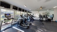Located at 99 Turtle Creek Dr in Asheville, NC, this well-appointed fitness center offers a comprehensive workout environment within a single open floor plan. The space is thoughtfully designed to accommodate a wide range of exercise needs, from cardiovascular training to strength and resistance work.

Large windows line one side of the room, flooding the space with natural light and offering pleasant views of the surrounding greenery — creating an energizing atmosphere for workouts. Floor-to-ceiling mirrors on the opposite wall visually expand the space and allow users to monitor their form throughout their sessions.

The cardio area features a row of elliptical trainers and stationary bikes, each equipped with individual monitor screens mounted above for entertainment or workout tracking. A dedicated strength training section houses a well-stocked Hampton dumbbell rack, medicine ball storage, a weight bench, and a multi-use cable machine, providing ample options for resistance training. Additional selectorized strength machines round out the equipment offerings.

Overhead fluorescent lighting ensures consistent illumination throughout, complemented by decorative ceiling fans. The durable rubber flooring is suited for high-traffic use. Health and wellness guidelines are posted on the walls, and convenient amenities such as a sanitizer dispenser and coat hooks are available near the entrance door, which opens to an outdoor parking area.