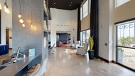 Located at Park West Green Drive in Katy, Texas, The Maddox presents a sophisticated and welcoming lobby space that immediately conveys a sense of modern elegance. The interior is defined by dramatic soaring ceilings that rise to impressive heights, creating an open and airy atmosphere enhanced by abundant natural light streaming through floor-to-ceiling windows dressed with floor-length charcoal drapery. Polished concrete floors run throughout, lending an industrial-chic aesthetic that anchors the contemporary design. The main reception area features a sleek, dark countertop workstation with quilted white chairs, ideal for leasing consultations. Adjacent to the entry, a stylish seating vignette showcases patterned wingback chairs arranged atop a cowhide rug, offering a welcoming first impression. Pendant Edison bulb lighting adds warmth and character to the space. A separate lounge area, finished with deep blue accent walls adorned with a curated gallery of framed artwork, provides an intimate retreat furnished with rich brown leather club chairs and a live-edge wood coffee table set over a bold crimson area rug. Glass-paneled doors connect the spaces, maintaining visual openness throughout. The branded entry doors offer clear sightlines to the surrounding Katy community.