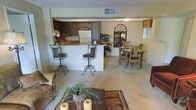 This single-floor space offers an open-concept design that effortlessly connects the living, dining, and kitchen areas into one cohesive, welcoming environment. The living room anchors the space with plush carpeting, a patterned area rug, and comfortable seating, creating an inviting atmosphere for relaxing or entertaining. A decorative coffee table adorned with candles and greenery adds a warm, homey touch.

The kitchen features dark wood cabinetry, a white refrigerator, and a raised breakfast bar with wrought iron barstools, providing both functionality and casual dining options. The bar counter serves as a natural visual divider between the kitchen and the living areas while maintaining the open feel of the floor plan.

Adjacent to the kitchen, the dining area accommodates a full dining table and chairs, making it ideal for shared meals. A decorative arched mirror on the wall adds an elegant focal point to this zone.

Throughout the space, neutral tones in the walls and flooring create a calm, cohesive backdrop, while warm lighting and thoughtful décor elements lend character and charm. The layout maximizes the sense of openness, making the space feel both functional and comfortable for daily living.