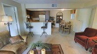 This single-floor space offers an open-concept design that effortlessly connects the living, dining, and kitchen areas into one cohesive, welcoming environment. The living room anchors the space with plush carpeting, a patterned area rug, and comfortable seating, creating an inviting atmosphere for relaxing or entertaining. A decorative coffee table adorned with candles and greenery adds a warm, homey touch.

The kitchen features dark wood cabinetry, a white refrigerator, and a raised breakfast bar with wrought iron barstools, providing both functionality and casual dining options. The bar counter serves as a natural visual divider between the kitchen and the living areas while maintaining the open feel of the floor plan.

Adjacent to the kitchen, the dining area accommodates a full dining table and chairs, making it ideal for shared meals. A decorative arched mirror on the wall adds an elegant focal point to this zone.

Throughout the space, neutral tones in the walls and flooring create a calm, cohesive backdrop, while warm lighting and thoughtful décor elements lend character and charm. The layout maximizes the sense of openness, making the space feel both functional and comfortable for daily living.