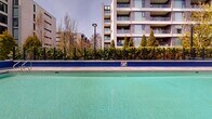 Located at 1515 W 57th Ave in Vancouver, BC, this outdoor aquatic facility offers a well-appointed recreational space set against a striking backdrop of classic brick architecture and contemporary residential towers. The centerpiece is a generously sized lap pool with clearly marked depth indicators, designed to accommodate both fitness swimming and leisure use. Its clear, turquoise water is enclosed by a broad, paved pool deck finished in neutral stone-toned pavers, providing ample space for lounging and movement around the water. Adjacent to the main pool sits a hot tub, offering a relaxing complement to the lap swimming experience. A dedicated information and signage kiosk is positioned near the pool entrance, providing facility rules and safety information for users. The perimeter of the facility is defined by sleek black fencing that frames the space while maintaining open sightlines to the surrounding greenery, including mature evergreen and deciduous trees that lend natural privacy and seasonal color. The facility benefits from open-sky exposure, allowing for abundant natural light throughout the day. The combination of the heritage-style brick building on one side and the modern multi-storey residential structure on the other gives this aquatic space a distinctive urban character, making it a notable amenity within the broader community.