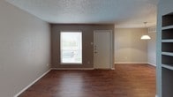 This single-floor unit is designed with a practical, straightforward layout finished in neutral tones throughout. The living area features rich, dark hardwood-style flooring and is illuminated by natural light streaming through a window fitted with horizontal blinds. A textured ceiling and clean white baseboards are consistent throughout the space. An open pass-through counter connects the living area to the kitchen, which is equipped with a coil-burner electric range, a full-size refrigerator, a dishwasher, and dark cabinetry complemented by tile backsplash and light hardwood-style flooring. Two carpeted bedrooms offer comfortable, quiet spaces, each with windows that allow natural light and neutral-toned walls. A ceiling fan with light fixture is installed in one bedroom. The bathroom features dark cabinetry with a white countertop, a wide vanity mirror with overhead lighting, and a combined tub and shower unit with white tile surround, paired with hardwood-style flooring. A second vanity area is accessible from the hallway. Closet access is visible along the corridor. The consistent use of gray-toned walls, textured ceilings, and a mix of flooring materials gives the unit a cohesive, clean aesthetic.