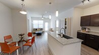This single-floor residence is designed with an open-concept layout that seamlessly connects the kitchen, dining, and living areas into one cohesive, light-filled space. The kitchen serves as a central focal point, featuring an expansive granite-topped island with an undermount sink and contemporary pendant lighting overhead. White cabinetry and clean finishes contribute to the modern aesthetic throughout.

Adjacent to the kitchen, a casual dining area accommodates a glass-top table with vibrant orange accent chairs, adding a pop of color to the otherwise neutral palette. The space transitions naturally into a generous living area, where large windows allow abundant natural light to pour in, enhancing the airy atmosphere. A ceiling fan with integrated lighting adds both function and comfort to the living space.

Hardwood-style flooring runs throughout, providing a warm, cohesive look. Louvered closet doors are visible near the living area, offering practical storage solutions. The overall design balances contemporary finishes with a welcoming, functional layout suited for everyday living.
