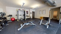 This single-floor fitness center is thoughtfully designed to support a comprehensive workout experience. The space is divided into distinct training zones, each outfitted with professional-grade equipment to accommodate a variety of exercise routines. The cardio area features multiple treadmills and is bathed in natural light from large windows that overlook an outdoor area, creating an open and energizing atmosphere. A floor-to-ceiling mirror lines one wall, enhancing the sense of space and allowing users to monitor their form during workouts. The strength training zone includes a multi-functional cable machine, adjustable weight benches, a full rack of dumbbells, medicine balls, and a plyo box — providing ample options for resistance and functional training. Workout reference charts are mounted on the walls for guidance. Durable rubber flooring covers the entire facility, offering both safety and comfort during high-impact activities. Exposed ductwork runs along the ceiling, lending the space an industrial aesthetic. A built-in water fountain and a waste receptacle are conveniently positioned near the entrance for ease of access. The facility is well-lit throughout with overhead fluorescent fixtures, ensuring a safe and functional environment for users at any time of day.