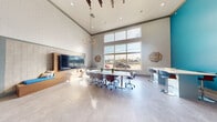 Located on West Dixie Highway in Miami, FL, this thoughtfully designed single-floor workspace offers a bright and contemporary environment ideal for productivity and collaboration. The space is defined by its dramatically high ceilings with exposed wooden beam accents, creating an open and airy atmosphere throughout. Large floor-to-ceiling windows flood the interior with natural light while providing views of the surrounding South Florida landscape, including swaying palm trees.

The main area features a central conference table with ergonomic rolling chairs, perfect for team meetings or group work sessions. A wall-mounted flat-screen TV set within a warm wood-paneled feature wall serves as a media and presentation hub, accompanied by a built-in bench seat with blue cushion accents. High-top bar tables along one side offer additional flexible seating for individual or collaborative work.

Near the entrance, built-in desk stations are positioned along wood-paneled walls, creating semi-private workspaces for focused tasks. A bold teal accent wall adds a vibrant splash of color to one section of the space, complementing the neutral gray tile flooring and light gray walls throughout. Decorative geometric wall art and a sculptural multi-arm chandelier add artistic character, rounding out this functional yet stylish Miami workspace.