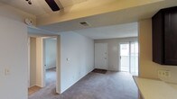 This single-floor residential space offers a functional and straightforward layout designed for comfortable everyday living. The main living area is finished with wall-to-wall carpet flooring, providing a soft and cohesive look throughout. A textured ceiling with a ceiling fan adds a classic touch to the space, while the smooth white walls create a bright, clean atmosphere.

The layout features an open flow between the living area and an adjacent room, visible through a wide doorway that hints at additional flexible living or sleeping space. The main entry is anchored by a solid white front door, complemented by a dark doormat that marks a clear transition point into the home.

Natural light plays an important role in the ambiance of this space, streaming in through a sliding glass door fitted with vertical blinds. This door likely leads to an outdoor area, offering both ventilation and connectivity to the exterior. A centrally placed ceiling vent ensures consistent climate control throughout the open living zone.

Overall, the space presents a clean, neutral palette with practical features that make it easy to personalize. The combination of carpeted flooring, ample natural light, and an open layout creates a welcoming and livable environment suited to a variety of lifestyles.
