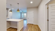 This single-floor unit showcases a thoughtfully designed open-concept layout with warm wood-look flooring flowing throughout the main living areas. The kitchen serves as a central highlight, featuring light gray shaker-style cabinetry, a white quartz countertop island, subway tile backsplash, stainless steel appliances, and a pair of elegant gold pendant lights overhead. Recessed lighting complements the bright, airy atmosphere throughout the space.

The living area extends naturally from the kitchen, offering ceiling fan fixtures and direct access to a private balcony through a glass-paneled door flanked by large windows dressed with white blinds, inviting ample natural light and outdoor views. The bedrooms are carpeted for comfort, each equipped with ceiling fans and generously sized windows that frame pleasant exterior scenery.

A dedicated laundry room accommodates a full-sized washer and dryer with overhead wire shelving for added storage. The bathroom is finished with a wide gray vanity, gold-toned fixtures, a framed mirror, and wood-look flooring that ties cohesively into the overall design palette. Clean, neutral tones and modern finishes throughout give the space a fresh, move-in-ready appeal.