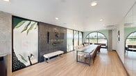 Located at 2603 North Fry Road in Katy, Texas, this contemporary meeting space delivers a polished and visually engaging environment designed for collaboration and gatherings. The single-floor layout makes efficient use of its footprint, centering around a sleek, elongated conference table surrounded by a mix of upholstered chairs that accommodate a generous number of attendees.

A defining architectural feature is the dramatic arched window at one end of the room, which frames outdoor views and floods the space with natural light. The smooth, light-toned hardwood flooring runs throughout, lending warmth and continuity to the interior. Recessed ceiling lighting supplements the natural light, ensuring a bright and focused atmosphere.

One wall is clad in a sophisticated dark textured tile, serving as a striking focal point and backdrop for a wall-mounted flat-screen display — ideal for presentations and media. Flanking this accent wall are frosted glass partitions adorned with artistic botanical illustrations, adding a creative, nature-inspired aesthetic while maintaining a sense of openness. Minimalist bench seating is positioned near the glass partitions and sliding glass doors, which open to an outdoor balcony offering additional views of the surrounding area.

The overall ambiance blends modern design sensibility with artistic detail, creating a versatile and inviting space suited for meetings, events, or collaborative work sessions.