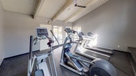 This fitness center is a purpose-built exercise facility designed to support a full cardio-focused workout experience. The space is defined by its distinctive vaulted ceiling with exposed wood-panel detailing and structural beams, lending the room an open, airy character. A ceiling fan is mounted at the apex, ensuring comfortable air circulation throughout workouts.

The facility is outfitted with multiple Precor USA treadmills arranged in a row along one wall, complemented by several elliptical cross-trainers positioned nearby. Large windows line the walls, inviting abundant natural light and offering views of the surrounding outdoor area, including what appears to be a courtyard or walkway. This connection to the exterior enhances the overall atmosphere of the space.

Full-length mirrors are installed along one wall, expanding the perceived depth of the room and allowing users to monitor their form during exercise. Motivational typography wall decals referencing strength, focus, and determination add a purposeful aesthetic to the gray-painted walls. Dark rubber flooring throughout provides durability and safety underfoot. A "Fitness Center" logo displayed prominently reinforces the dedicated nature of this well-maintained amenity space.