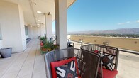 Located on Lankershim Boulevard in North Hollywood, this rooftop terrace delivers a striking open-air experience defined by its commanding elevation and unobstructed panoramic views. The space stretches generously along the upper level of the building, featuring large-format tile flooring throughout and a clean, contemporary architectural design with structural columns supporting a partial overhead covering that provides shade along one side of the terrace.

The covered section of the terrace is furnished with woven rattan lounge chairs accented by bold red and patterned throw pillows, creating inviting seating arrangements ideal for relaxed gatherings. Additional dining-style seating is positioned in a more open area, accompanied by potted tropical and ornamental plants that add a touch of greenery to the urban setting. Large floor-to-ceiling aluminum-framed windows line the interior-facing wall, allowing natural light to flow both inward and outward while reflecting the mountain scenery.

The open terrace area extends beyond the covered zone, featuring a glass and metal railing system that preserves sightlines across the sprawling San Fernando Valley and toward the rugged San Gabriel Mountains. The abundant Southern California sunlight, open skies, and far-reaching mountain backdrop make this rooftop terrace a standout feature of the property.