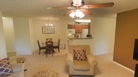 This single-floor residence features an open-concept layout that seamlessly connects the living, dining, and kitchen areas into one cohesive and functional space. The living area is anchored by a ceiling fan with integrated lighting, providing both comfort and illumination, while a decorative chandelier adds an elegant touch above the dining zone.

The kitchen is positioned at the far end of the open floor plan, separated by a raised breakfast bar with a granite-style countertop that doubles as a casual seating or serving area. Medium-toned wood cabinetry lines the kitchen walls, complemented by white appliances including a refrigerator and microwave. The design allows for easy interaction between the kitchen and the main living areas.

The dining area accommodates a round table with seating for four, positioned beneath the chandelier and framed by a framed botanical wall print. Soft, neutral-toned carpet extends throughout the space, contributing to a warm and inviting atmosphere. The overall color palette of creams and warm beiges creates a cohesive, comfortable environment well-suited for everyday living and casual entertaining.