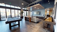 This contemporary amenity lounge is designed to impress, combining industrial and modern design elements across a single, open floor plan. Soaring ceilings with exposed ductwork and structural beams lend the space an urban loft character, while a slatted wood ceiling canopy over the lounge zone adds warmth and definition. Large-format stone tile flooring runs throughout, grounding the space with a sleek, durable finish.

The game area is anchored by a full-size pool table and a shuffleboard table, positioned beneath stylish pendant lighting that adds focused illumination. Adjacent to the game zone, a well-appointed wet bar features a mosaic tile backsplash, open floating shelves, dark cabinetry, and bar-height seating — ideal for social gatherings. A live-edge communal dining table nearby adds a natural, artisanal touch.

The lounge area offers plush sectional seating arranged around a wall-mounted flat-screen TV, complemented by bold abstract artwork and designer area rugs. Floor-to-ceiling black-framed glass doors and clerestory windows flood the interior with natural light and provide seamless visual and physical access to a spacious outdoor patio, where additional seating overlooks the surrounding community. The overall ambiance is sophisticated, social, and inviting.