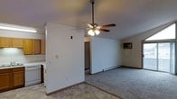 This single-floor space features an open-concept layout that connects the kitchen and living areas in a functional, straightforward design. The kitchen, positioned to one side, includes light wood cabinetry, a white range, and tile flooring that provides a practical surface for food preparation and daily use. A partial wall divides the kitchen from the living area, creating a subtle sense of separation while maintaining the open feel of the space.

The living area is carpeted and offers a comfortable, understated environment. A ceiling fan with a built-in light fixture is centrally mounted, providing both air circulation and ambient lighting. Climate control is further supported by a baseboard heating unit along one wall and a wall-mounted air conditioning unit, ensuring year-round temperature regulation.

The walls are neutral white throughout, offering a clean, versatile backdrop. A hallway visible through a doorway suggests additional rooms or storage beyond the main living space. The overall layout is practical and efficient, suited for everyday living with clearly defined functional zones within a compact, single-level footprint.