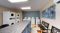 This shared laundry facility is designed for convenience and functionality, offering residents a practical and organized space to handle their laundry needs. The room features a row of top-loading washing machines along the back wall, complemented by a set of stacked dryers along the side wall, providing ample capacity for multiple users at once. A large, laminate-topped folding counter runs along one side of the room, offering generous surface space for sorting and folding laundry. The floor is dressed in a distinctive multicolored tile pattern featuring orange, yellow, and white geometric shapes, adding a lively visual element to the utilitarian space. A window with horizontal blinds along the far wall allows natural light to fill the room while offering a view of the greenery outside. The walls are painted in a clean combination of light yellow and a muted steel blue accent, contributing to a calm and neutral atmosphere. Overhead fluorescent lighting ensures the space remains bright and well-lit at all times. A pair of wrought iron benches provides comfortable seating for those waiting on their laundry. The overall layout is efficient and user-friendly, making this laundry room a highly functional shared amenity.