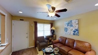 This single-floor space presents a warm and welcoming living area defined by its neutral yellow-toned walls and textured ceiling. The room is centered around a ceiling fan with an integrated light fixture, providing both airflow and ambient illumination. Natural light enters through a window dressed with dark curtain panels and horizontal blinds, contributing to a balanced and comfortable atmosphere.

The living area is furnished with a rich brown leather sofa and a coordinating patterned armchair, arranged around a dark wood coffee table that anchors the seating space. A decorative multi-drawer chest serves as a functional accent piece beneath the window, topped with a traditional table lamp that adds a warm glow. A large floral canvas artwork in shades of blue and white adds a decorative focal point to one wall, introducing a touch of color to the otherwise earthy palette.

Carpeted flooring throughout the living area enhances the cozy feel of the space. The front entry door is positioned to one side, keeping the layout practical and accessible. Overall, the space offers a comfortable and neatly arranged environment with thoughtful decorative touches.
