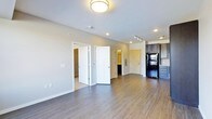 This single-floor space offers a thoughtfully designed open layout that promotes a sense of spaciousness and easy flow between living areas. The main living area is finished with rich, dark hardwood-style flooring that contrasts beautifully against the light gray walls and white trim, lending the space a clean, modern aesthetic. Recessed lighting and a central flush-mount ceiling fixture provide ample illumination throughout the open area, while a track lighting system near the kitchen adds focused task lighting.

The kitchen is positioned to one side and features dark cabinetry with a sleek finish, paired with a black refrigerator that adds a bold, contemporary touch. The cabinetry offers generous storage, and the kitchen layout is designed for functional use.

Multiple doorways lead from the main living area to additional rooms, including what appears to be a bedroom and a walk-in closet or storage area, with a glimpse of a bathroom visible in the background. The doors feature classic paneled designs with white finishes, complementing the overall neutral color palette.

Throughout the space, clean lines, consistent flooring, and a cohesive color scheme create a calm, inviting ambiance that is both practical and visually appealing.