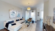This single-floor residence presents a thoughtfully designed living area centered around an open-concept layout that maximizes both comfort and functionality. The living room serves as the focal point of the space, featuring hardwood-style flooring in a cool gray tone that runs throughout, complementing the crisp white walls and neutral color palette.

A drum pendant light fixture hangs from the smooth ceiling, providing warm ambient lighting that pairs with the generous natural light streaming through large floor-to-ceiling windows dressed in sheer white curtains. The windows offer views to an outdoor balcony area, enhancing the sense of openness and connection to the exterior.

The living area is well-proportioned, accommodating a sectional seating arrangement alongside accent furniture with ease. Framed geometric artwork adorns the walls, adding visual interest without overwhelming the clean aesthetic. A mirrored surface is visible in an adjacent area, further amplifying the bright, airy ambiance. The overall design language is contemporary and cohesive, emphasizing light tones, modern fixtures, and an uncluttered arrangement that makes the space feel welcoming and versatile.