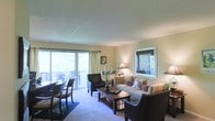 This single-floor space centers around a well-appointed living room that balances comfort and functionality. Large sliding glass doors along one wall flood the interior with natural light and provide direct access to an outdoor deck, where glimpses of lush surrounding trees can be seen. A secondary window on the adjacent wall further enhances the bright, airy atmosphere throughout the room.

The living area is finished with neutral-toned carpet and warm beige walls, creating a welcoming and cohesive color palette. Furnishings include a generously sized sofa with decorative pillows, complemented by dark-framed accent chairs arranged to encourage conversation. A wooden coffee table anchors the seating arrangement, while a table lamp adds warm ambient lighting. Decorative touches such as a framed orchid arrangement and wall art contribute to the polished, livable character of the space.

Light-colored sheer curtains frame the sliding doors, softening the natural light while maintaining the connection to the outdoors. Overall, the living space offers a comfortable and inviting environment with a strong emphasis on natural light and outdoor integration.