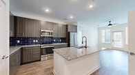 This well-appointed 2-bedroom, 1-bathroom residence showcases a modern design aesthetic with high-quality finishes throughout its single-level floor plan. The open-concept kitchen and living area serves as the heart of the home, where a striking kitchen commands attention with gray shaker-style cabinetry, bold navy blue subway tile backsplash, granite countertops, and a full suite of stainless steel appliances. A large center island with a deep undermount sink and designer faucet provides additional prep space and casual seating. Light wood-tone flooring runs throughout the main living areas, complementing the bright, neutral palette. The living room is bathed in natural light from large windows and a glass door leading to a private balcony, with a ceiling fan adding comfort overhead. Both bedrooms feature plush carpeting and ceiling fans, with the generously sized primary bedroom offering access to a walk-in closet equipped with built-in shelving. The bathroom is elegantly finished with dark hexagonal tile flooring, granite dual-sink vanity, and a tiled tub-shower combination with decorative stripe accents. A second walk-in closet provides ample additional storage throughout the home.