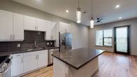 Located at 1818 Park Ave in Dallas, TX, this single-floor unit offers a sleek, modern design with thoughtful finishes throughout. The open-concept kitchen is a standout feature, equipped with stainless steel appliances including a French door refrigerator and dishwasher, crisp white cabinetry, a dark tile backsplash, and a central island with a dark countertop ideal for meal prep or casual dining. Pendant lighting above the island adds a contemporary touch. Warm, light-toned hardwood flooring flows seamlessly throughout the living areas, complementing the neutral gray walls and creating a cohesive, inviting ambiance. The living area is generously proportioned, featuring a ceiling fan and a large window fitted with horizontal blinds that invite natural light while maintaining privacy. An additional room, also finished with hardwood floors and a bright window, offers flexible use as a bedroom or office. The bathroom is elegantly appointed with large-format marble-look tile on both the floors and walls, a frameless glass-enclosed walk-in shower with hexagonal mosaic tile flooring, a floating vanity with a dark countertop, and a well-lit mirror. Recessed lighting throughout the unit enhances the clean, contemporary aesthetic of this well-appointed Dallas residence.