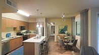 This single-floor space showcases an open-concept layout that integrates the kitchen, dining, and living areas into one cohesive environment. The kitchen is well-appointed with warm wood cabinetry, stainless steel appliances — including a full-size refrigerator and microwave — and a sleek white-topped island with bar stool seating, ideal for casual dining or meal prep. Two pendant lights hang above the island, complementing the track lighting overhead to create layered illumination throughout the space.

Hardwood-style flooring runs continuously across the entire area, adding warmth and visual continuity. The dining area, situated adjacent to the kitchen, features a round glass-top table surrounded by contemporary upholstered chairs, with a bold green abstract artwork anchoring the nearby wall. A large window dressed with floor-length drapes allows natural light to filter into the space.

A hallway visible from the main area leads toward additional rooms, suggesting a thoughtful and functional floor plan. The overall ambiance is modern and welcoming, combining practical design with tasteful finishing touches throughout.