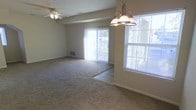 This single-floor space offers an open and functional layout designed for comfortable everyday living. The main living and dining area is generously sized, featuring plush wall-to-wall carpeting that transitions seamlessly into tile flooring near the sliding glass door — a practical and visually appealing design detail. Large windows fitted with horizontal blinds allow abundant natural light to fill the space, creating a bright and welcoming atmosphere throughout the day. A full-length sliding glass door provides direct access to an outdoor area and offers views of the exterior surroundings, enhancing the sense of openness. The dining area is defined by an elegant multi-bulb chandelier with a classic bronze finish, adding a touch of traditional charm. The living area is complemented by a ceiling fan, ensuring year-round comfort. Neutral-toned walls and beige carpeting create a clean, versatile backdrop that accommodates a wide range of interior styles. A floor vent is visible along the wall, indicating a central HVAC system for climate control. Overall, the space presents a straightforward, move-in-ready environment with practical finishes and a layout that maximizes usable living area.