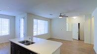 This single-floor space showcases an open-concept layout that seamlessly connects the kitchen and living areas. The kitchen features a white countertop island with a double stainless steel sink, providing both functionality and a clean aesthetic. Dark cabinetry beneath the island creates a striking contrast against the light countertop surfaces.

The living area is bathed in natural light, thanks to a large window fitted with horizontal blinds and an adjacent glass door, allowing for ample brightness throughout the day. Smooth, light-toned walls complement the warm wood-look flooring that extends across the entire space, adding continuity and warmth.

A ceiling fan with a built-in light fixture is centrally positioned in the living area, offering both air circulation and supplemental lighting. A partial wall with a pass-through opening adds architectural interest while maintaining the open feel of the layout. The overall ambiance is clean, neutral, and move-in ready, with a minimalist design that provides a versatile backdrop for a variety of interior styles.
