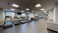 This fitness center is a comprehensively equipped, single-floor facility designed to support a full range of exercise needs. The space is organized into distinct functional zones, allowing for efficient use of the floor area throughout.

The cardio zone features a row of treadmills and elliptical machines positioned along one wall, beneath a wall-mounted flat-screen television. Ceiling-mounted dual fans circulate air throughout this section, contributing to a comfortable workout environment. Horizontal windows along the lower portion of the walls allow natural light to filter in while maintaining privacy.

The strength training area includes multiple Cybex cable and resistance machines, free-weight dumbbells on a dedicated rack, and stability equipment such as exercise balls and balance discs. Full-length mirrors line portions of the walls, enhancing the sense of space and allowing users to monitor their form during exercise.

Throughout the facility, durable speckled rubber flooring provides traction and impact absorption. A drop ceiling with recessed panel lighting ensures consistent, even illumination across all areas. A separate entry vestibule features a wall-mounted television, a water station, and accessible restroom facilities, serving as a functional transition area between the entrance and the main workout floor.
