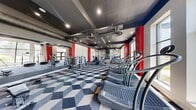 This fitness center offers a spacious, single-floor layout designed to accommodate a comprehensive range of workout activities. The interior is defined by a striking industrial aesthetic, with exposed ductwork and recessed lighting set against a dark ceiling, creating a modern and energetic atmosphere. Bold red accent columns punctuate the white walls, adding visual contrast throughout the space.

The flooring features a distinctive geometric cube-pattern carpet in shades of blue, grey, and white, lending a dynamic visual element underfoot. Large windows along the perimeter allow natural light to fill the space while offering views of the surrounding exterior.

The cardio zone is well-stocked with Woodway treadmills, Cybex ellipticals, stair climbers, and stationary bikes, including upright and recumbent options by Spirit. A dedicated strength training area houses a multi-station cable machine by True Fitness, dumbbell racks, and weight benches, with full-length mirrors enhancing visibility and spatial perception.

A standout design feature is the large, colorful abstract mural that serves as a focal point near the cardio section, adding an artistic and vibrant energy to the environment. A wall-mounted television and additional mirrors are also present, rounding out a functional and visually engaging fitness facility.
