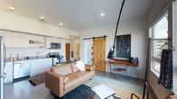 This single-floor space showcases a thoughtfully designed open-concept layout that integrates the living area and kitchen into one cohesive environment. The living area is anchored by a tufted tan leather sofa positioned atop layered area rugs on polished concrete floors, lending a warm yet modern aesthetic. A mid-century-inspired workspace with a wooden desk and an articulating pendant lamp occupies one corner, accompanied by a wire accent chair and a striking piece of abstract wall art, adding visual depth to the space.

A standout architectural feature is the natural knotted wood sliding barn door, which provides both a rustic contrast and a functional room divider. The kitchen, visible from the living area, features white cabinetry, stainless steel appliances including a microwave and range, and a clean, efficient layout. Recessed ceiling lighting throughout ensures consistent, even illumination across the space. The neutral white walls and concrete flooring create a versatile backdrop that balances the warmth of the wood accents and leather furnishings, resulting in a cohesive, contemporary atmosphere well-suited for both relaxing and working.
