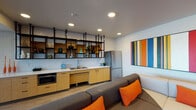 This contemporary amenity lounge is thoughtfully designed to offer a dynamic social environment. The open-plan layout seamlessly connects multiple functional zones, beginning with a well-appointed kitchenette. Warm bamboo cabinetry paired with crisp white countertops and a stainless steel undermount sink set an inviting tone, while a striking open black metal shelving system above the subway tile backsplash displays curated decorative vessels and greenery. A stainless steel refrigerator and built-in microwave complete the space.

The lounge area centers around a generously sized sectional sofa accented with vibrant orange throw pillows, anchored beneath a bold large-format striped canvas artwork on a clean white wall. Recessed lighting throughout maintains a bright, welcoming atmosphere.

Adjacent to the lounge, a games area features a full-size billiards table positioned beneath pendant and track lighting. This zone flows directly into a dramatic floor-to-ceiling glass wall that opens to a spacious outdoor terrace, flooding the interior with natural light and offering sweeping panoramic views of the surrounding landscape. A warm wood-paneled accent wall provides a natural counterpoint, complete with a large wall-mounted flat-screen television, creating an ideal media viewing area atop vibrant orange carpet tile flooring.