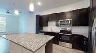 This modern single-floor unit offers a thoughtfully designed layout with contemporary finishes throughout. The kitchen serves as a centerpiece, showcasing rich dark cabinetry that reaches toward the ceiling, granite countertops with a striking salt-and-pepper pattern, and a full suite of stainless steel appliances including a range, microwave, dishwasher, and refrigerator. A decorative geometric tile backsplash adds visual interest, while a large granite-topped island provides additional prep space and casual seating. Pendant lighting overhead enhances the kitchen's modern aesthetic. Light wood-look flooring flows throughout the main living areas, lending warmth and continuity to the open-concept floor plan, which connects the kitchen seamlessly to the living space. The primary bathroom features a double-sink vanity topped with matching granite, dark cabinetry with sleek hardware, large mirrors, and a glass-enclosed walk-in shower with tiled surround, as well as an adjacent walk-in closet. The bedrooms offer plush carpeting and natural light through tall windows with blinds, while a ceiling fan in the primary bedroom ensures year-round comfort. Clean white walls and recessed lighting throughout contribute to a bright, contemporary ambiance.