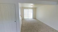 This two-floor residence presents a straightforward and functional design with clean, neutral finishes throughout. The main living area, visible in the image, is a generously sized room finished with wall-to-wall beige carpeting that lends a warm, understated feel to the space. Cream-colored walls and white trim create a bright, cohesive aesthetic that carries throughout the interior.

A key feature of the main living area is the large sliding glass door, which serves as both a source of natural light and a direct connection to the outdoors. The door provides views of surrounding greenery, bringing a sense of openness to the room. A flush-mount ceiling light fixture provides additional illumination, keeping the space well-lit regardless of the time of day.

A partial wall division with an open archway defines separate zones within the living area, adding subtle architectural interest without compromising the open feel. A white paneled door is also visible, likely leading to a closet or adjoining room. The overall ambiance is calm and neutral, offering a blank canvas that accommodates a variety of interior styles. The two-floor layout further provides additional living space and functional separation between areas of the home.