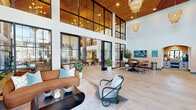 This expansive amenity facility offers a thoughtfully designed, single-floor layout encompassing a diverse range of functional and social spaces. The grand entry lobby immediately impresses with soaring double-height ceilings, floor-to-ceiling black-framed glass walls, warm wood slat ceiling accents, and woven rattan pendant chandeliers. Plush seating arrangements and large-format tile flooring create a welcoming, resort-inspired atmosphere flooded with natural light.

The coworking lounge features floor-to-ceiling windows overlooking the surrounding neighborhood, providing an abundance of natural light. Rows of long wooden workstations with acoustic privacy dividers and distinctive slatted pendant lighting make it ideal for focused productivity.

The entertainment lounge is appointed with a bold blue tile accent wall, a large wall-mounted display, bar-height seating, and a billiards table atop a vibrant, patterned area rug — perfect for social gatherings.

The kitchen area showcases rich walnut cabinetry, glossy terracotta-toned tile backsplash, concrete countertops, and a matte black undermount sink, delivering a refined, contemporary aesthetic.

The fitness center is generously proportioned with high ceilings, bamboo-style flooring, and a full complement of Life Fitness strength and cardio equipment, including a functional training rig and a heavy bag. Arched architectural detailing and a staircase complete the well-connected layout.