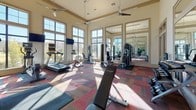 This fitness center is designed to support a comprehensive workout experience within a spacious, well-appointed environment. The facility boasts soaring ceilings accented by wood crown molding and equipped with multiple ceiling fans to maintain air circulation throughout. Large, floor-to-ceiling windows line the walls, flooding the interior with abundant natural light and offering outdoor views of the surrounding community. The colorful patchwork carpet flooring adds a dynamic visual element to the space.

The cardio zone is positioned along the windowed wall and features treadmills, elliptical machines, and spinning bikes, each equipped with individual monitors. Wall-mounted flat-screen televisions provide additional entertainment options during workouts. The strength training area includes a variety of cable and resistance machines suitable for full-body conditioning, along with a dedicated free weight section featuring a multi-tier dumbbell rack. An adjustable workout bench supports a range of strength exercises.

Full-length wall mirrors span a significant portion of the interior, enhancing the sense of openness and allowing users to monitor their form. The layout is thoughtfully organized to provide ample space between equipment, ensuring a comfortable and functional workout environment. Glass-paned double doors contribute to the bright, airy atmosphere of this well-maintained fitness facility.