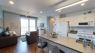 This contemporary single-floor unit offers a thoughtfully designed open-concept layout that seamlessly integrates the kitchen, dining, and living areas. The kitchen is outfitted with white cabinetry, white appliances, and a spacious peninsula countertop with seating — ideal for casual dining or entertaining. Pendant and track lighting illuminate the space efficiently throughout.

The living area features warm hardwood flooring, a muted blue accent wall, and an exposed concrete ceiling that lends an urban, industrial character to the space. A large flat-screen TV is mounted above a streamlined media console, while floor-to-ceiling sliding glass doors flood the room with natural light and open to sweeping views of the surrounding cityscape and distant mountain range.

The bedroom is a serene retreat, carpeted for comfort, with a corner window that offers elevated, wide-open views of the city below and clear skies beyond. The bathrooms are finished to a high standard: one features a sleek, all-white glass-enclosed walk-in shower with white subway tile walls and chrome fixtures, while the second bathroom includes a long vanity with white cabinetry, a wide mirror, and wood-look tile flooring. Together, these spaces offer a clean, modern aesthetic with a cohesive palette of whites, grays, and natural wood tones.