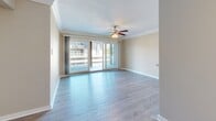 This single-floor residence showcases a cohesive, well-maintained interior with a neutral color palette and thoughtful finishes throughout. The main living area is bright and open, featuring light-toned hardwood-style flooring, crisp crown molding, and a ceiling fan with a built-in light fixture. A large sliding glass door floods the space with natural light and provides direct access to a balcony, offering views of the surrounding exterior. The textured ceiling adds a classic residential touch, while clean white baseboards frame the room neatly.

A secondary room, likely a bedroom, continues the crown molding detail and features plush wall-to-wall carpeting for added comfort. A ceiling fan mirrors the style found in the living area, and a window allows natural light to filter in. The warm beige walls create a calm, inviting atmosphere throughout both rooms.

The galley-style kitchen is efficiently laid out with gray shaker cabinetry extending to the upper and lower walls on both sides. Marble-look countertops provide a polished surface alongside a stainless steel double sink. Black appliances, including a refrigerator and electric range, complement the contemporary cabinetry, while light hardwood-style flooring and crown molding carry the design language consistently into this functional space.