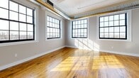 Located at 2075 Kenmore Ave in Buffalo, NY, this 1-bedroom, 1-bathroom loft-style residence blends industrial character with modern finishes. The open-concept kitchen and living area serves as the heart of the space, showcasing soaring ceilings with exposed silver ductwork and structural piping that lend an authentic industrial aesthetic. The kitchen is outfitted with crisp white cabinetry, a subway tile backsplash, stainless steel appliances, and a marble-topped center island illuminated by a trio of bold navy and gold pendant lights. Rich hardwood floors run throughout, adding warmth against the bright white walls. A long hallway connects the main living area to the private bedroom, where large, multi-pane windows framed in black flood the room with natural light and offer sweeping views of the Buffalo skyline. Exposed ductwork continues into the bedroom, reinforcing the loft-style character. The bathroom features a full tub-shower combination with clean white tile surround and built-in shelving. Industrial-style cage pendant lights in the hallway complete the cohesive design theme, creating a space that is both functional and architecturally distinctive.