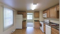 This two-floor space includes a well-equipped kitchen that serves as a central feature of the home. The kitchen is outfitted with warm-toned wood cabinetry arranged along both walls, providing ample storage through upper and lower cabinet configurations. Dark laminate countertops contrast the lighter cabinetry, adding visual depth to the space. Appliances include a full-sized white refrigerator, a gas range with oven, and a built-in microwave, offering a complete cooking setup. The flooring throughout the kitchen is light hardwood, contributing to a clean and open aesthetic. A flush-mounted fluorescent light fixture on the textured ceiling provides bright, even illumination. A window at the far end of the kitchen allows natural light to filter into the space, with a view of greenery outside. An open doorway leads from the kitchen into an adjacent carpeted room, suggesting a connected layout that promotes flow between living areas. The overall design of the kitchen is practical and straightforward, with a neutral color palette of beige and cream tones creating a cohesive, understated atmosphere throughout.