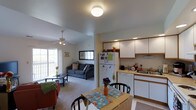 This single-floor unit presents an open-concept layout that seamlessly connects the living, dining, and kitchen areas into one cohesive space. The living area is carpeted and benefits from natural light streaming through a sliding glass door that leads to a balcony. A ceiling fan with a light fixture keeps the space comfortable, while a sofa and coffee table create a cozy seating arrangement. An adjacent dining area accommodates a wooden table with seating for multiple guests, accented by a decorative centerpiece. The kitchen is efficiently organized with white upper and lower cabinetry offering ample storage, laminate countertops, and a full suite of appliances including a refrigerator, dishwasher, and sink positioned beneath recessed cabinetry lighting. The transition from carpeted living areas to hard flooring in the kitchen is clearly defined. Flush-mount ceiling lighting illuminates the central areas of the unit. The overall layout is practical and well-suited for everyday living, with each zone flowing naturally into the next within a modest, functional footprint.