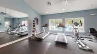 This fitness center is a thoughtfully designed, single-floor facility offering a comprehensive workout environment across several distinct zones. The space is finished with cool blue-gray walls, white trim, and durable carpet tile flooring throughout, creating a clean and motivating atmosphere.

The main cardio area is anchored by multiple treadmills and an elliptical machine, positioned in front of large windows that overlook an outdoor pool, flooding the room with natural light and providing pleasant views during workouts. Full-length mirrors line one wall, visually expanding the space and assisting with form checks. A rack of medicine balls and a wall-mounted exercise guide add functional detail to this zone.

Adjacent to the cardio area, a dedicated strength training section houses a multi-station cable and weight machine, supported by instructional exercise charts mounted on the wall. A water fountain is conveniently located nearby. The vaulted ceiling in this area adds a sense of openness and volume.

A separate, quieter room serves as a yoga and stretching studio, complete with pink yoga mats, a stability ball, a decorative potted plant, and natural light from a large window. Workout reference posters adorn the walls, making it an ideal space for mindfulness or cool-down routines.

An additional room provides space for functional training, featuring a power tower for bodyweight exercises, a kettlebell rack, and a wall-mounted television. Windows on two sides bring in ample natural light and views of the surrounding landscape.