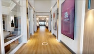 This 3,960-square-foot single-floor amenity space is thoughtfully designed to offer residents a comprehensive lifestyle experience. The centerpiece is a spacious clubroom with light hardwood flooring, tray ceilings, and recessed lighting. It features a large sectional sofa, a wall-mounted TV set against a bold blue accent wall, and a full kitchen bar with leather counter stools — ideal for social gatherings. Adjacent to the lounge is a game area equipped with a shuffleboard table, framed by an expansive panoramic window overlooking an outdoor balcony and courtyard. The fitness center is a standout feature, spanning a generous area with a bold herringbone-patterned carpet. It is outfitted with cardio machines including treadmills, strength training equipment such as a power rack and cable machines, free weights, medicine balls, and large wall mirrors that amplify the sense of space. Natural light filters in through multiple windows. A stylish corridor with modern sconce lighting, circular pendant fixtures, plush lounge chairs, and curated artwork connects the various areas. Additional amenities include glass-door private offices or meeting rooms and a convenience store, all finished with consistent light hardwood flooring throughout.