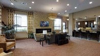 This single-floor space presents a well-appointed communal lounge area designed with both comfort and style in mind. The focal point of the room is a striking stacked-stone accent wall that houses a sleek linear fireplace and a wall-mounted flat-screen television, creating an inviting atmosphere for gathering and relaxation. The space is generously lit by a combination of recessed ceiling lights and a bold square pendant light fixture that adds a modern touch overhead. Large windows dressed with patterned, floor-length drapes line the walls, allowing natural light to fill the room while offering a view of the surrounding exterior. The seating arrangement is thoughtfully curated, featuring neutral-toned sofas and accent chairs paired with dark-framed coffee tables and cube ottomans, offering flexible and comfortable seating options. The warm-toned carpeting grounds the space and contributes to its welcoming ambiance. Adjacent to the main lounge area, a connected kitchenette or bar counter with bar seating is visible, adding a functional element to the overall layout. The combination of textured stone, warm neutrals, and contemporary furnishings gives this space a polished yet approachable character, suitable for socializing or quiet leisure.
