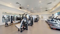 Located in Buckeye, Arizona, this fitness center offers a generously sized, single-floor exercise room designed to support a comprehensive workout experience. The space features a clean, modern aesthetic with crisp white walls, smooth neutral-toned flooring, and recessed lighting complemented by multiple ceiling fans that promote airflow throughout the room.

The strength training area is well-appointed with a variety of commercial-grade resistance machines, a multi-station cable system, and a dedicated free weights section featuring a full dumbbell rack displayed in front of a wall-length mirror — ideal for form monitoring and maximizing the sense of space. The cardio zone includes treadmills, stair climbers, stationary spin bikes with individual monitors, and a rowing machine, with wall-mounted flat-screen TVs positioned for easy viewing during workouts.

Natural light streams in generously through large, dark-framed windows along multiple walls, creating a bright and energizing atmosphere. Entry points include both a set of double glass doors and a single glass door, providing easy access. Additional amenities include a waste station and wall-mounted control panels for climate and media. The open, uncluttered layout ensures ample room to move freely between equipment zones, making this fitness center functional, inviting, and well-suited for a wide range of training routines.