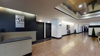 This single-floor space presents a polished, contemporary design well-suited for a professional or commercial environment. The layout flows from a reception area through extended corridors and into a thoughtfully appointed lounge, creating a cohesive and functional sequence of spaces.

The entry and corridor areas are defined by speckled terrazzo tile flooring complemented by dark border detailing, which contrasts sharply against crisp white walls. Matte black doors punctuate the hallways, reinforcing the clean, modern aesthetic. Recessed and wall-mounted lighting illuminates the passages evenly, while decorative wall art and a slim console table add subtle character along the corridor's length. Potted topiary plants introduce organic texture throughout.

The lounge area serves as the visual centerpiece of the space. A layered ceiling with perimeter cove lighting draws the eye upward, creating a sense of vertical dimension. Furnishings include a channel-tufted grey sofa, deep rust-toned accent chairs with patterned cushions, and a wooden bench with leather upholstery, all arranged around a round coffee table atop a neutral area rug. A framed abstract artwork anchors one wall, while a vertical green hedge panel adds a natural, design-forward element. Glass entry doors allow views to the exterior, inviting natural light into the reception zone.